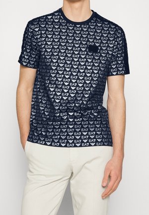 Mann trägt ein dunkelblaues T-Shirt mit sich wiederholenden weißen EA7-Logos und beigen Hosen, steht mit einer Hand in der Tasche.
