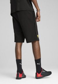 Pantalones cortos negros con un logotipo amarillo de Ferrari, que cuentan con una franja lateral con texto. Llevados con calcetines negros y zapatillas deportivas con detalles rojos.