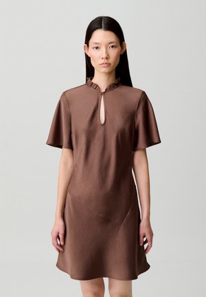 Femme aux longs cheveux foncés portant une robe marron à manches courtes avec un décolleté en trou de serrure et un col à volants, debout devant un fond uni.