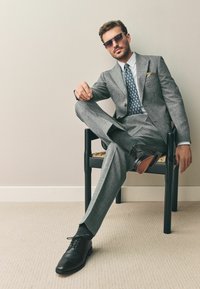 Hallgrey ülikond mustrirohkega lipsuga ja tasku ruuduga, mustad nahkkingad, istudes mustas toolis, heledal taustal, tekstuuriga vaip.