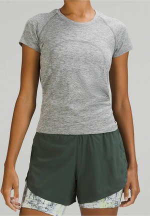 Frau trägt ein eng anliegendes graues kurzärmeliges Sportoberteil und dunkelgrüne, mehrlagige Laufshorts mit gemusterten Innenshorts.