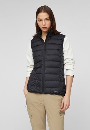 Donna con indosso un gilet nero trapuntato senza maniche sopra una camicia bianca a maniche lunghe e pantaloni cargo beige su uno sfondo grigio semplice.