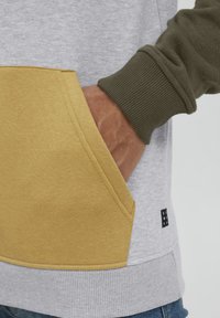 Grauer Sweatshirt mit einer großen gelben Tasche und grünen Rippbündchen. Der Stoff wirkt strukturiert, mit einem Markenetikett, das am Saum aufgenäht ist.