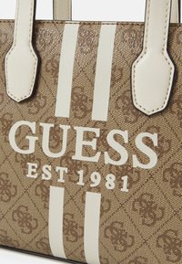 Sac à main marron à motifs avec accents crème, arborant le logo "GUESS EST 1981" et deux rayures verticales. Texture lisse, matériau durable.