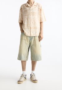 Beige korte mouw shirt met geometrische patronen, gecombineerd met lichte denim shorts. Grijs-witte sneakers met marineblauwe accenten maken de look compleet.