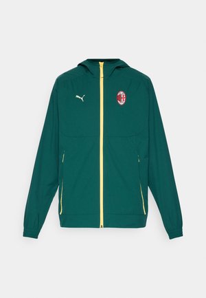 Grøn letvægtsjakke med lynlåslukning, gule detaljer, hætte og et fremtrædende rødt og sort klublogo på venstre bryst.