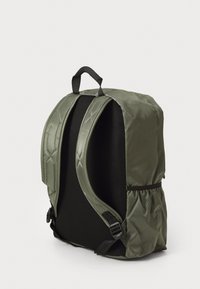 Mochila verde con respaldo de malla negra acolchada, correas de hombro ajustables, bolsillo elástico lateral y asa de transporte en la parte superior, sobre un fondo liso.