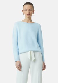 Hellblauer Pullover aus geripptem Strick mit weitem Ausschnitt und langen Ärmeln, kombiniert mit weißen Hosen, die eine Bindeschnalle in der Taille haben.
