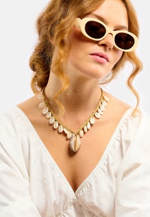 Sonnenbrille in Cremefarbe, mehrschichtige Muschel-Halskette mit einer zentralen Kaurimuschel, schlichtes weißes Oberteil mit strukturierter Stoffstruktur. Lockiges Haar fällt sanft.