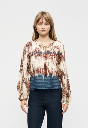 Młoda kobieta ubrana w luźną bluzkę z długim rękawem o geometrycznym wzorze w kolorach brązowym, kremowym i niebieskim, do tego ciemnoniebieskie dżinsy.