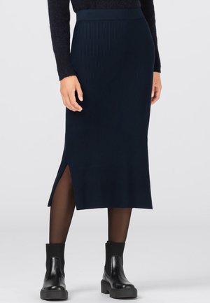 Kvinde iført en marineblå ribbet midi nederdel med sideslids, sorte gennemsigtige strømpebukser, sorte ankelstøvler og en sort, fluffy sweater.