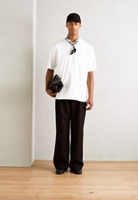 Vit oversized t-shirt med bröstficka, svarta vida byxor, svart keps och mönstrad scarf. Håller en svart väska.