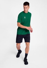 Grøn bomulds t-shirt med korte ærmer og en logo, kombineret med sorte shorts. Modellen bærer sorte sko og sokker og går på en ensfarvet baggrund.