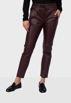 Personne portant un pantalon jogging en cuir bordeaux foncé avec une taille à cordon et des chaussures noires slip-on, debout devant un fond clair uni.