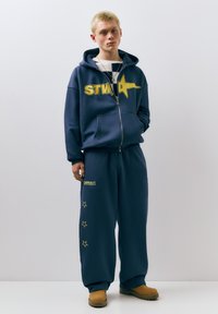 Young person with short blond hair wearing navy blue hoodie and pants with yellow star designs, brown boots, standing against plain background.
