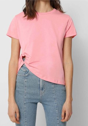 Femme portant un t-shirt rose clair à manches courtes avec un détail froncé sur le côté, associé à un jean en denim bleu taille haute.