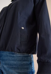 Gros plan d'une personne portant une veste bleu marine avec une poche à pression et un jean en denim bleu.