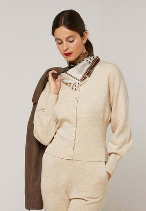 Femme en cardigan beige boutonné et pantalon tenant une veste marron sur l'épaule, portant un foulard en soie à motifs et un rouge à lèvres rouge.