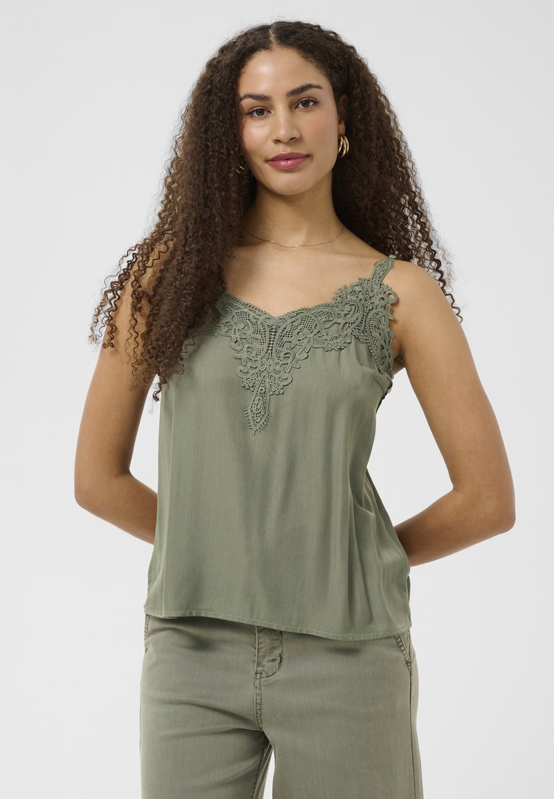 Jeune femme aux longs cheveux bouclés portant un haut vert sans manches avec un détail en dentelle et un pantalon assorti, debout les mains dans le dos.
