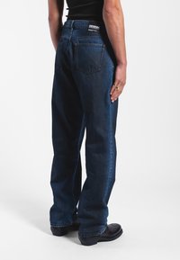 Tmavě modré džínové kalhoty s volným střihem, viditelným švem, dvěma zadními kapsami a štítkem na pase. Nohavice doplněné černými botami.