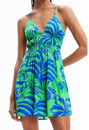 Vrouw draagt een mouwloze, groene en blauwe mini-jurk met tropische print, dunne schouderbandjes en een diepe V-hals.