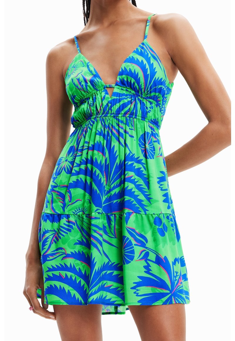 Femme portant une mini-robe sans manches à imprimé tropical vert et bleu avec des fines bretelles et un décolleté en V profond.