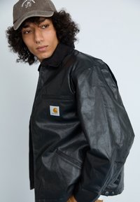 Giubbotto di pelle nera con una tasca con zip e un'etichetta con il logo Carhartt. Il colletto è in velluto a coste nero e il tessuto ha una texture leggermente lucida.