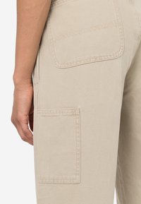 Beige cargobyxor i strukturerat tyg, med två stora fästrappar på sidorna och detaljerad sömnad runt kanterna.