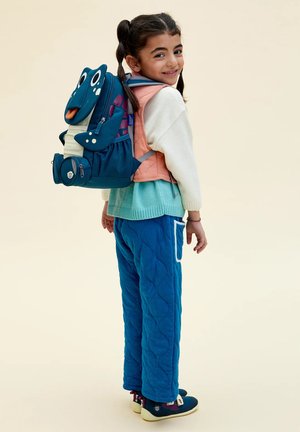 Smiling girl with pigtails wears pastel sweater, blue quilted pants, and a playful blue dog-shaped backpack, standing against a light background.