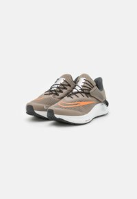 Nike-sneakers met een bruin en oranje ontwerp, met een gestructureerde bovenkant, zwarte veters en een witte, gewatteerde zool met een gemusterde buitenzool.