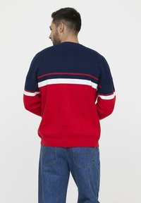 Pull rayé rouge et marine avec des accents blancs, col et poignets côtelés. Porté avec un jean en denim bleu, affichant une coupe décontractée.
