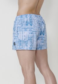 Mėlynos raštuotos maudymosi šortai su elastinga juosta. Audinys pasižymi geometriniais raštais, kurių atspalviai svyruoja nuo mėlynos iki baltos.