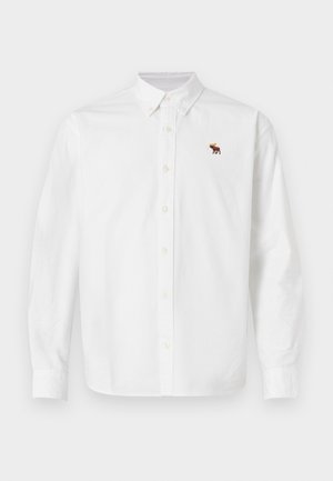 Chemise blanche boutonnée en coton. Elle comprend des manches longues, un col classique et une petite broderie d'orignal sur le côté gauche de la poitrine.