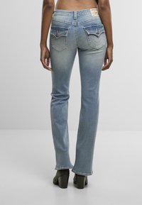 Asmuo, vilkintis šviesiai mėlynas bootcut džinsus ir juodus aukštakulnius batus, stovi prieš baltą fono.