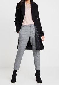 Cappotto nero lungo su un top rosa chiaro, abbinato a pantaloni a sigaretta grigi a quadri e stivali neri alla caviglia, che mettono in mostra una silhouette elegante e aderente.