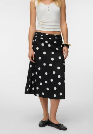 Femme portant un haut blanc sans manches, une jupe midi noire à pois blancs, des ballerines noires et des bracelets à son poignet.