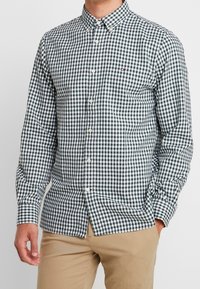 Camisa de cuadros verde y blanca, de botones, con cuello, mangas largas y un logo de marca en el pecho, hecha de tela ligera.