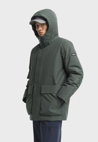 Zelená zateplená bunda s kapucňou, predným zipsom, veľkými vreckami a logom na rukáve. Hladká textura a moderný dizajn.