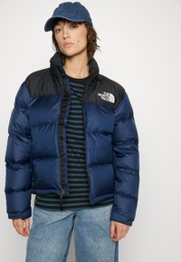 Marineblå puffjakke med sorte skuldre, med høy krage, glidelås foran og logodetaljer. Bært over en stripete skjorte og jeans.
