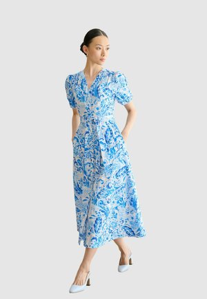 Frau mit dunklem Haar in einem niedrigen Dutt, trägt ein blau-weißes, floral gemustertes Midikleid mit Puffärmeln und weißen Absätzen, geht mit den Händen in den Taschen.