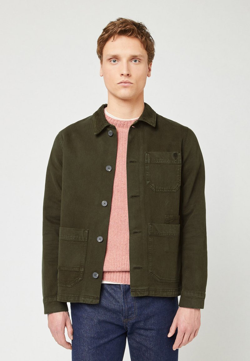 Olive green jacket with a collar, button front, two front pockets, and soft texture. Worn over a light pink sweater. Simple design.
