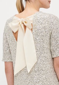 Glittrande klänning i guld med korta ärmar och öppen rygg, med en stor krämfärgad rosett vid halsen. Slät textur, glänsande finish.