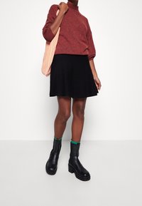 Persona che indossa un maglione color ruggine, una gonna nera, stivaletti neri alla caviglia con calze verdi e che porta una borsa a tracolla a righe arancioni e bianche.