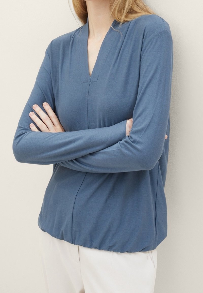 Sininen pitkähihainen paita, jossa V-pääntie, valmistettu pehmeästä kankaasta, rento malli ja koottu helma. Sujuva rakenne, minimaalinen muotoilu.