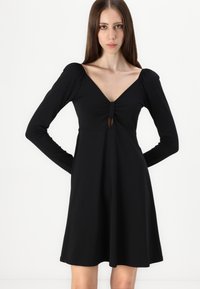 Veste preta com mangas longas, decote em V e detalhe central em forma de buraco. Tecido suave, saia rodada e detalhes franzidos nos ombros.