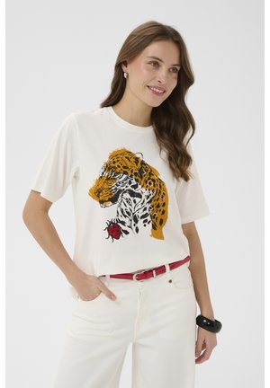 Žena u bijeloj majici s velikim printom leoparda i dizajnom crvenog srca, bijelim hlačama, crvenim remenom i crnom narukvicom, blago se smiješi.