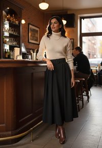 Kremowy sweter z golfem, czarna spódnica z wysokim stanem i plisami, brązowe botki za kostkę, stojąca przy drewnianym barze w dobrze oświetlonej kawiarni.