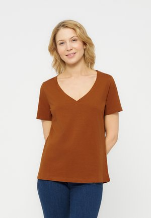 Kobieta z blond włosami, ubrana w brązową koszulkę z krótkim rękawem i dekoltem w serek oraz niebieskie dżinsy, stojąca na tle jednolitego jasnego tła.