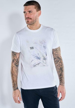 Man with tattoos on both arms wearing white Emilio Adani T-shirt and dark pants, looking to his left against a plain light background.