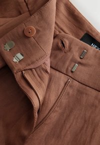 Zbliżenie na brązowy materiał ubrania, pokazujące guzik, dziurkę na guzik, zamek błyskawiczny oraz metalowe zapięcia typu hak i oczko na pasku od spodni.